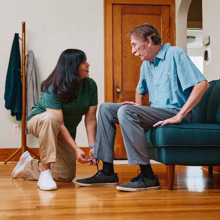 A Home Instead caregiver kneels to help an elderly man tie his shoes in a cozy living room, highlighting patient and personalized care for daily tasks.