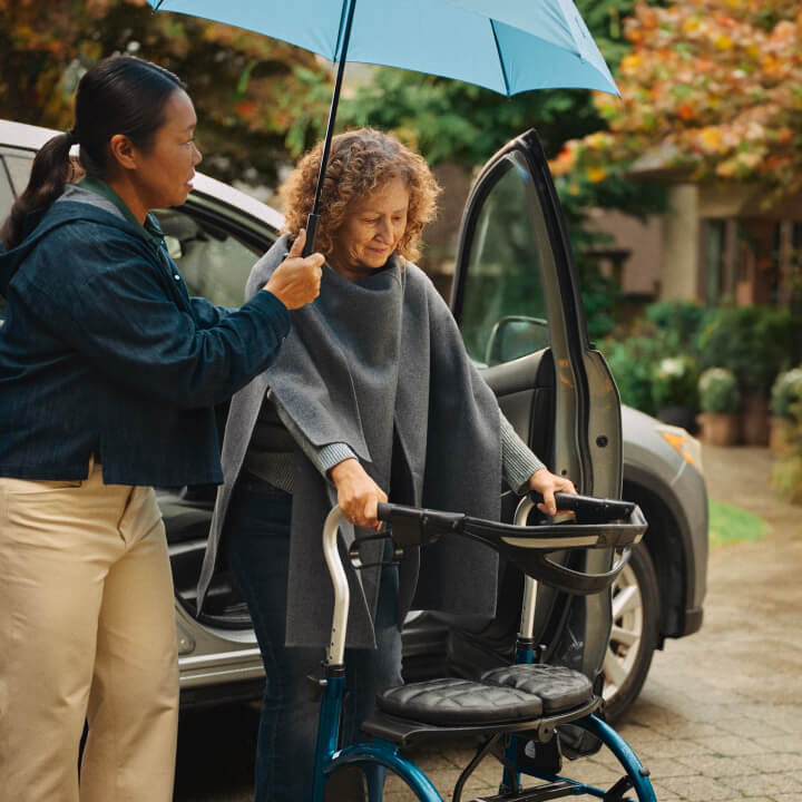 A Home Instead caregiver holds an umbrella for an elderly woman using a walker as they step out of a car on a rainy day, highlighting attentive and thoughtful care.