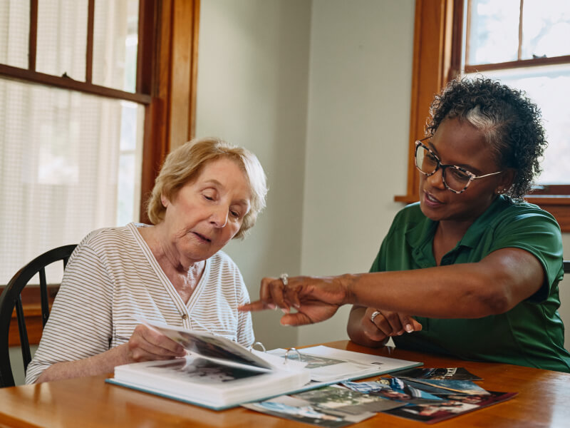 A Home Instead caregiver and an elderly woman review a photo album together at a dining table, sharing memories and stories.