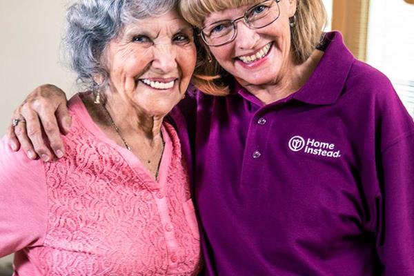 Home Instead Caregiver and senior woman with cane stand smiling together at home.